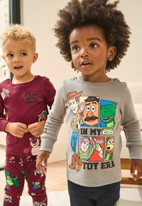 Dwoje dzieci w tematycznych ubraniach: jedno w bordowej koszulce z długim rękawem z motywem Toy Story, a drugie w szarej koszulce z kolorowymi grafikami postaci.