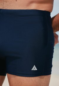 Marinefarbene Badehosen aus glattem Material verfügen über ein weißes Logo auf der unteren linken Seite. Der Schnitt reicht hoch über den Oberschenkel und sitzt eng.