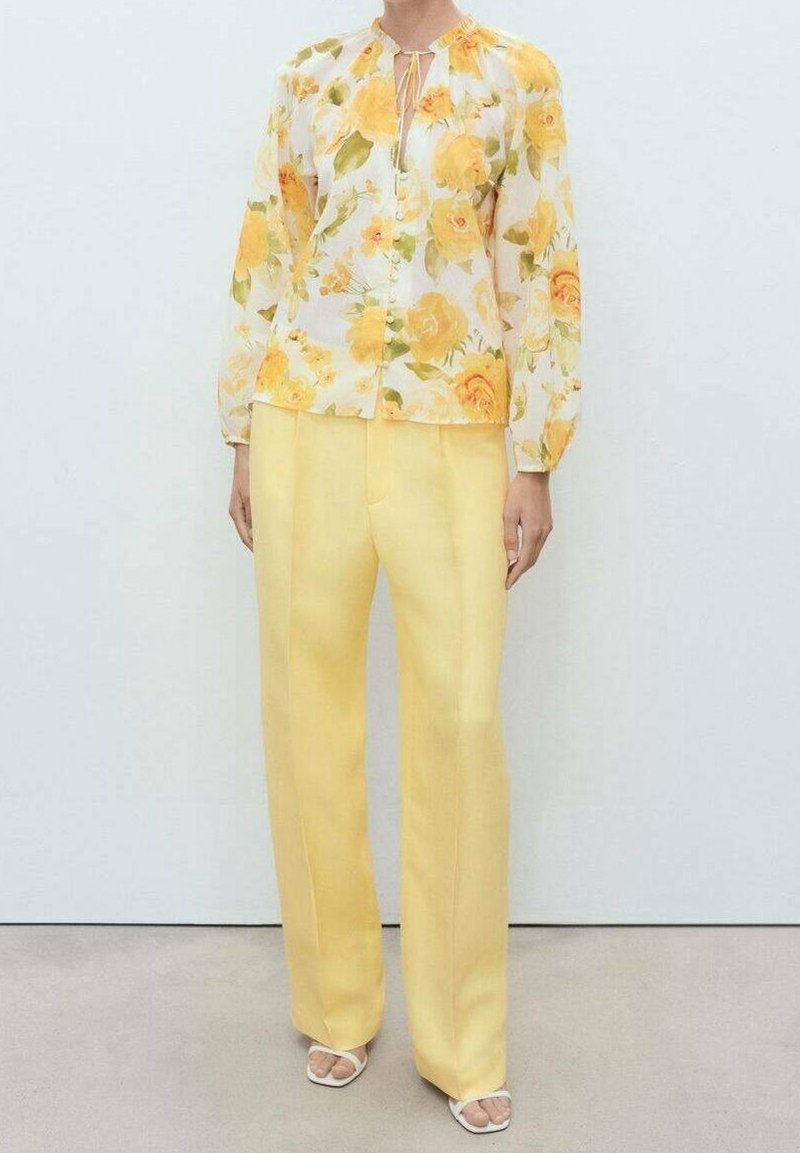 Personne portant un chemisier blanc à imprimé floral jaune et un pantalon ample jaune clair, debout sur un sol neutre contre un mur blanc.