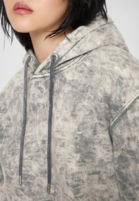 Sweat à capuche dans un motif gris délavé et crème, avec des cordons gris et un tissu doux et texturé. Coupe décontractée, sans logos visibles.