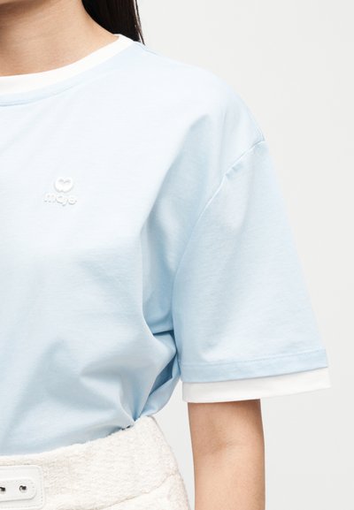 Άτομο που φοράει ανοιχτό γαλάζιο πουκάμισο με κοντά μανίκια, με λευκή λεπτομέρεια σε στρώσεις στα μανίκια και ένα μικρό λογότυπο στο στήθος.