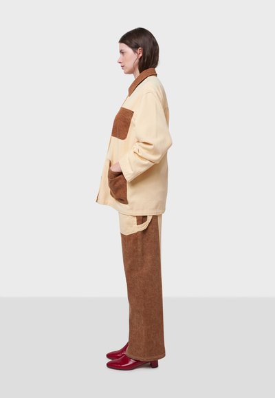 Beige kordjacka med bruna accenter och fickor, kombinerad med bruna byxor. Röda klackar fullbordar outfiten, uppvisad mot en grå bakgrund.