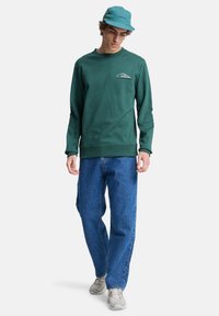 Sudadera verde con cuello acanalado, con un logotipo blanco en el pecho. Combinada con unos vaqueros azules holgados y zapatillas grises.