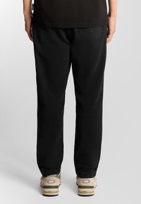 Zwarte sweatpants met een relaxte pasvorm, voorzien van een achterzak en een gladde textuur. Gepaard met beige sneakers en een zwart shirt.