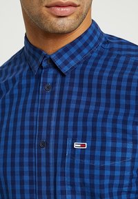 Niebieska koszula w kratę zapinana na guziki, z ciemnymi i jasnoniebieskimi kratkami, spiczastym kołnierzykiem, kieszenią na piersi oraz wyszywanym logo.