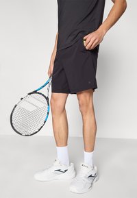 Črne atletske kratke hlače z žepom, v katerem je teniška žogica, obute v bele čevlje in z ročajem teniške loparice z modrimi poudarki.