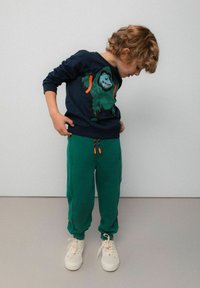 Ciemnoniebieska bluza z grafiką zielonego goryla, zestawiona z zielonymi spodniami dresowymi z pomarańczowymi sznurkami oraz białymi trampkami.