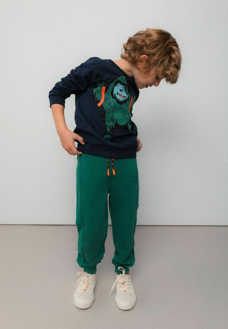 Ciemnoniebieska bluza z grafiką zielonego goryla, zestawiona z zielonymi spodniami dresowymi z pomarańczowymi sznurkami oraz białymi trampkami.