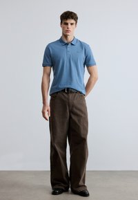 Gaiši zils polo krekls ar apkaklīti, kombinēts ar brūnām plaša piegriezuma biksēm. Modelis stāv pret neitrālu fona sienu, valkājot melnus apavus.