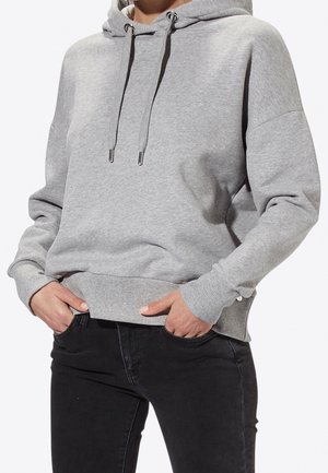 Person trägt hellgrauen Kapuzenpullover mit Kordeln und schwarze Jeans, Hände in den vorderen Taschen, Oberkörper gezeigt.