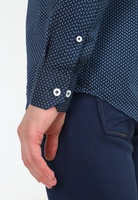 Navy blue shirt with white dot pattern, button cuffs, and smooth cotton fabric. Hand resting alongside dark blue pants.