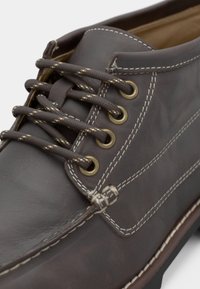 Brune lærsko med rund tå, kontrasterende hvit søm og messingøyler. Har teksturerte snører og en flat gummisåle.