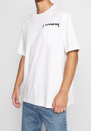 Mann trägt ein weißes Lacoste-T-Shirt mit Brusttasche und blaue Jeans, steht vor einem schlichten Hintergrund.
