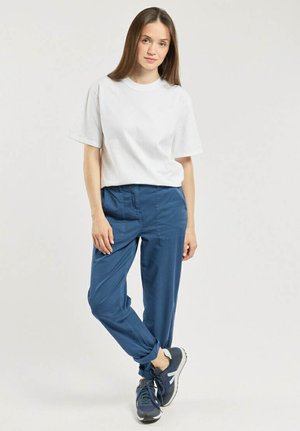 Jeune femme debout, jambes croisées, portant un t-shirt blanc à manches courtes, un pantalon bleu et des baskets bleues sur un fond uni.