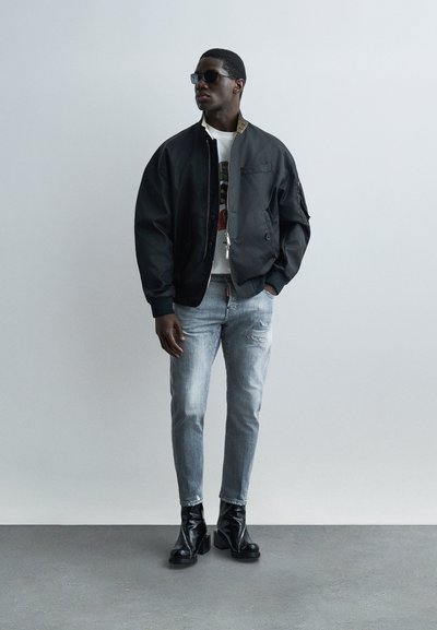 Homme portant une veste bomber noire, un t-shirt blanc à motif, un jean clair déchiré, des bottines à talons noires et des lunettes de soleil, debout contre un mur gris uni.