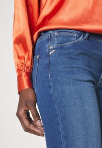 Personne portant un chemisier en satin orange vif et un jean en denim bleu, main détendue le long du corps sur un fond neutre.