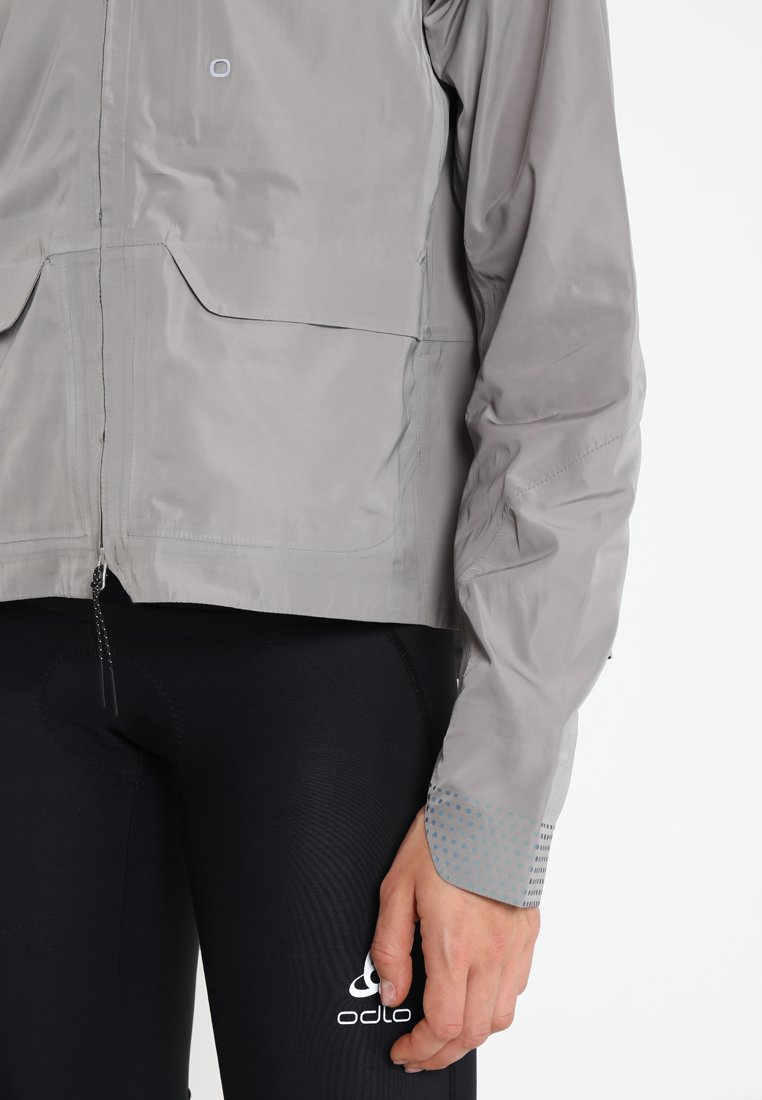 Hellgraue sportliche Jacke mit glatter Textur, Fronttaschen und Polka-Dots-Akzenten an den Bündchen. Trägt sie über eng sitzenden schwarzen Leggings.