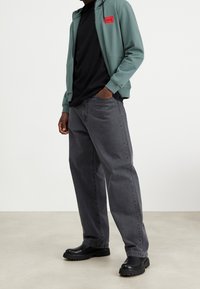 Osoba stojící na jednolité podlaze, oblečená v zelené mikině na zip s červenou nášivkou, černém tričku, volných šedých džínách a černých robustních botách.