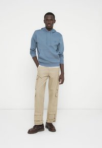 Helehall sinise kapuutsiga dressipluus esitaskuga, kombineeritud beežide mitme taskuga cargo-pükstega ja tumepruunide paeltega kingadega.