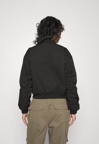 Černá bomber bunda s žebrovaným límcem a manžetami, šikmými švy a nařasenými rukávy, v kombinaci se světle béžovými cargo kalhotami.