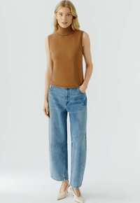 Bruine mouwloze gebreide turtleneck gecombineerd met lichtblauwe wijde jeans. De outfit wordt afgerond met crème hoge hakken.