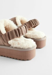 Des chaussons en platform marron avec une doublure moelleuse crème et une sangle froncée. Extérieur en daim avec une semelle texturée en zigzag. Apparence douce et confortable.