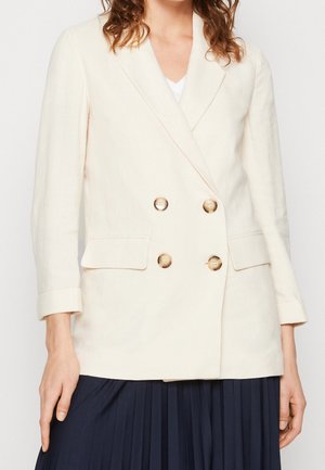 Kobieta nosząca kremową dwurzędową marynarkę z czterema guzikami i kieszeniami z patkami na białą bluzkę i ciemną plisowaną spódnicę.