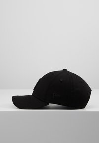 Casquette de baseball noire à visière courbée, vue de côté, présentée sur une surface blanche contre un fond gris clair uni.