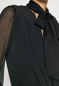 Black blouse with sheer, billowy sleeves and a knotted necktie detail. Features a draped silhouette and smooth fabric texture.