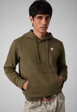 Giovane uomo con felpa verde oliva con tasca frontale e pantaloni chiari con fantasia, in piedi davanti a uno sfondo grigio uniforme.