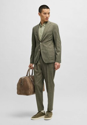 Olijfgroen pak gemaakt van gestructureerde stof met twee zakken, gecombineerd met een lichtgroen shirt. Accessoires zijn een bruine tas en sneakers.
