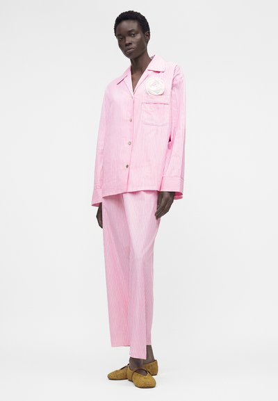 Μοντέλο φοράει άνετο ροζ και λευκό ριγέ τοπ στυλ πιτζάμας με λευκό λουλούδι στην τσέπη και ταιριαστό φαρδύ παντελόνι με υφαντά παπούτσια.