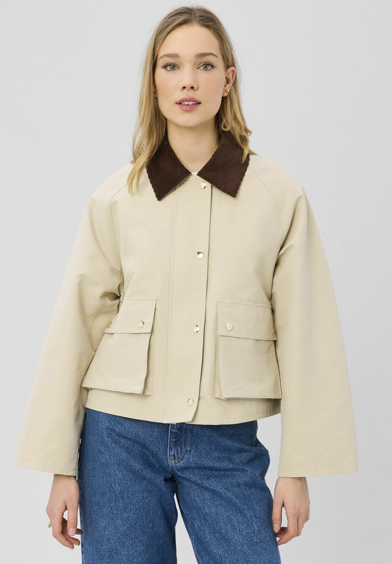 Jeune femme portant une veste beige avec un col marron et de grandes poches frontales, associée à un jean bleu, debout devant un fond uni.