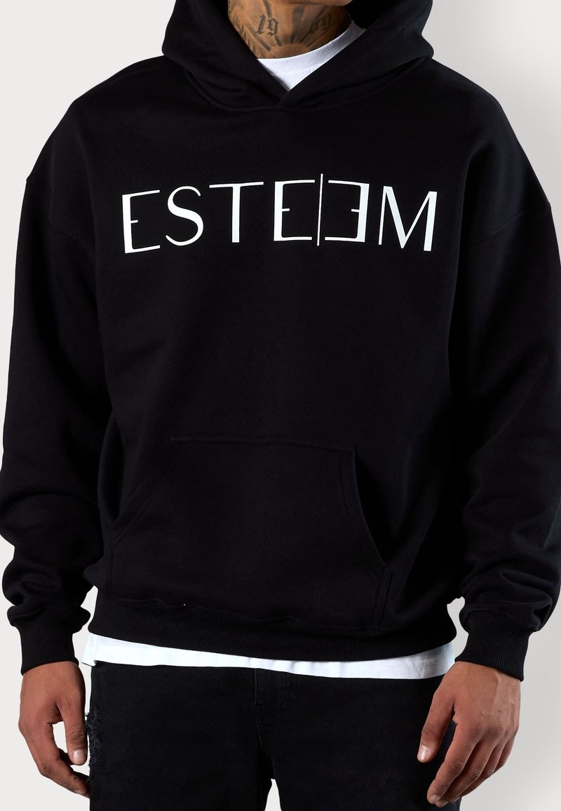 Sweat à capuche noir avec poche avant et texte stylisé blanc "ESTEEM", porté sur une chemise blanche, tatouage partiellement visible sur le cou du porteur.