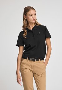 Polo noir avec col, manches courtes, patte de boutonnage à trois boutons, et un petit logo blanc sur la poitrine, associé à un pantalon kaki.