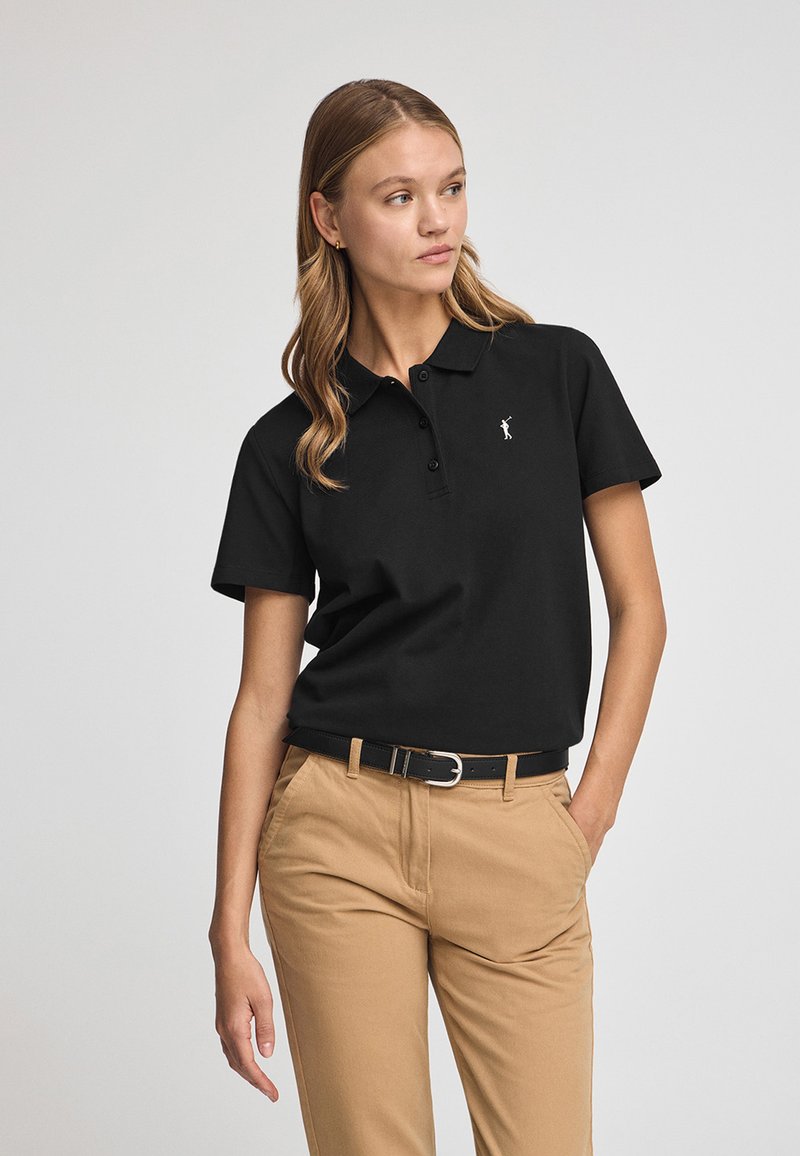 Polo noir avec col, manches courtes, patte de boutonnage à trois boutons, et un petit logo blanc sur la poitrine, associé à un pantalon kaki.