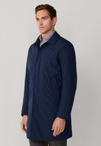 Man in een marineblauwe lange jas over een gestreept shirt, staand tegen een effen lichtgrijze achtergrond.