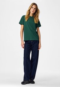 Jonge vrouw met lang blond haar, gekleed in een donkergroen T-shirt met korte mouwen, losse donkerblauwe jeans en zwarte sneakers, staand tegen een witte achtergrond.