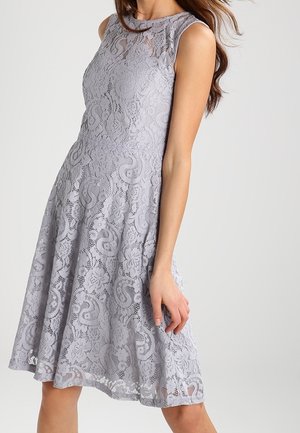 Robe sans manches en dentelle lavande longueur genou avec motifs floraux et tourbillons, corsage ajusté et jupe évasée portée par une femme.