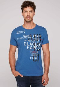 Camiseta de algodón azul con cuello redondo, mangas cortas y acentos de texto gráfico en blanco y rojo. Textura suave, corte casual.