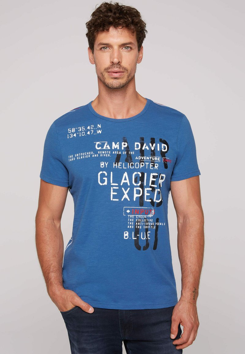 Camiseta de algodón azul con cuello redondo, mangas cortas y acentos de texto gráfico en blanco y rojo. Textura suave, corte casual.