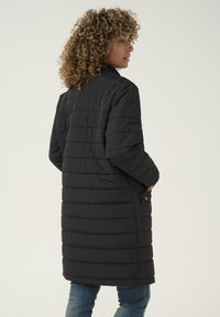 Abrigo de plumas negro con un diseño largo y acolchado, cuello acanalado y bolsillos laterales, mostrado desde la parte de atrás.