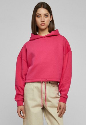 Rosa cropped Hoodie mit Kordelzug und übergroßen Ärmeln, aus weichem Material. Gestylt mit beigen Hosen, vor einem neutralen Hintergrund.