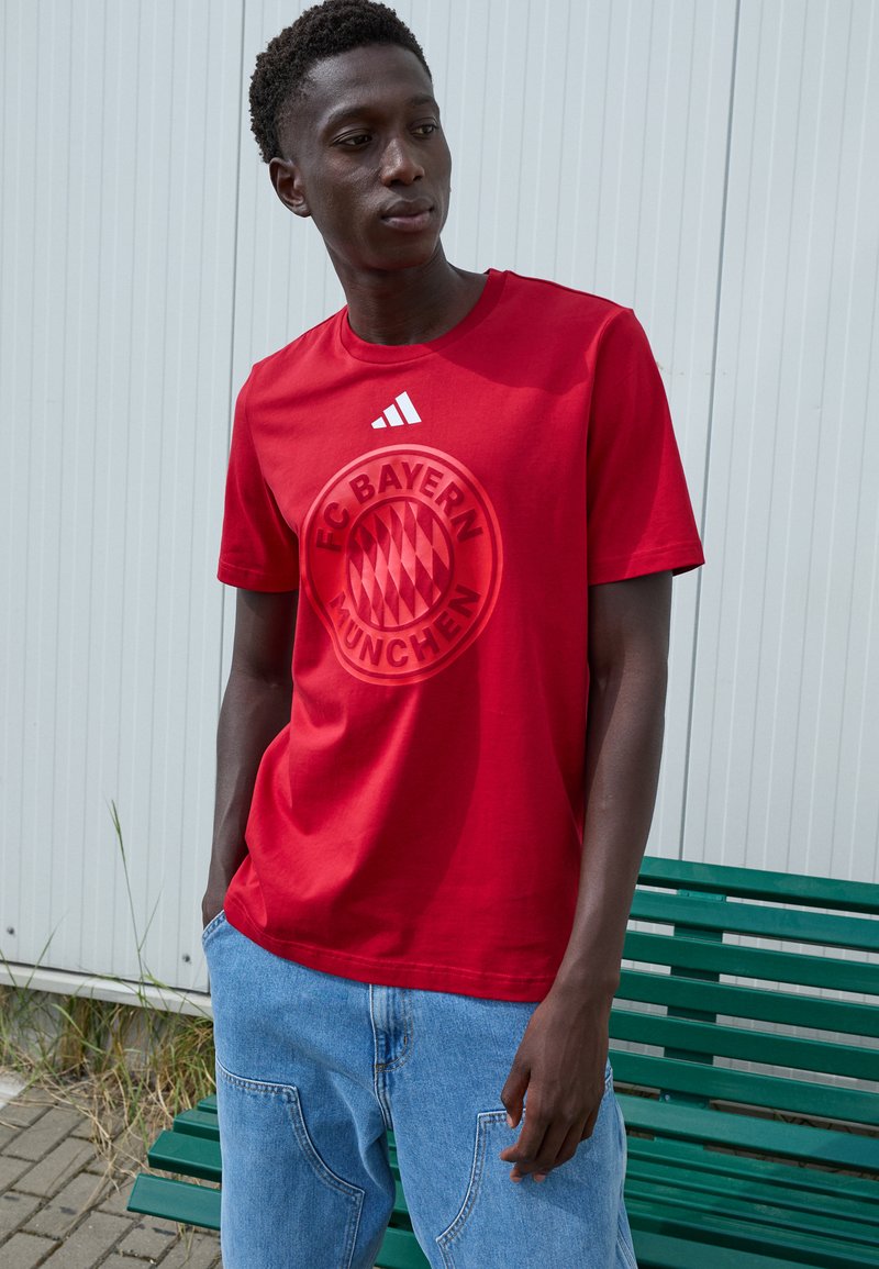 Red cotton t-shirt featuring a raised FC Bayern Munchen logo on the front, with white adidas stripes on the neckline. Blue denim shorts.