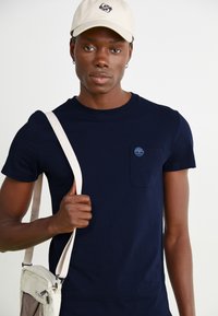 Tricou din bumbac bleumarin, cu gât rotund, mâneci scurte și o buzunare pe piept cu un logo albastru. Geantă crossbody bej cu curea ajustabilă.