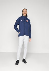 Navy zip-up jacket with hood, featuring a pink Nike logo and team emblem, paired with white slim-fit track pants and black athletic shoes.