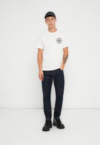 Tricou alb din bumbac cu un logo imprimat negru pe piept, purtat împreună cu blugi denim de culoare bleu închis și bocanci negri cu șiret.