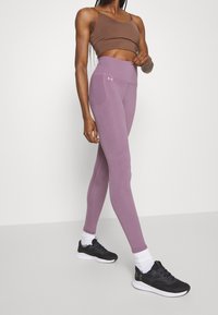 Legginsy w jasnym fioletowym kolorze z wysokim stanem i boczną kieszenią, w połączeniu z brązowym crop topem oraz czarnymi butami do biegania w białych skarpetkach.