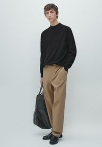 Tmavý pletený rolák, béžové plisované kalhoty, černé kožené boty, držení velké černé tašky přes rameno. Minimalistický design, neutrální barvy.