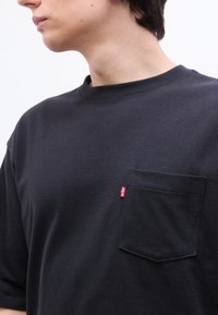 Κοντινό πλάνο ενός ατόμου που φοράει μαύρο μπλουζάκι με στρογγυλή λαιμόκοψη, με τσέπη στο στήθος και μια μικρή κόκκινη ετικέτα Levi's στη άκρη της τσέπης.